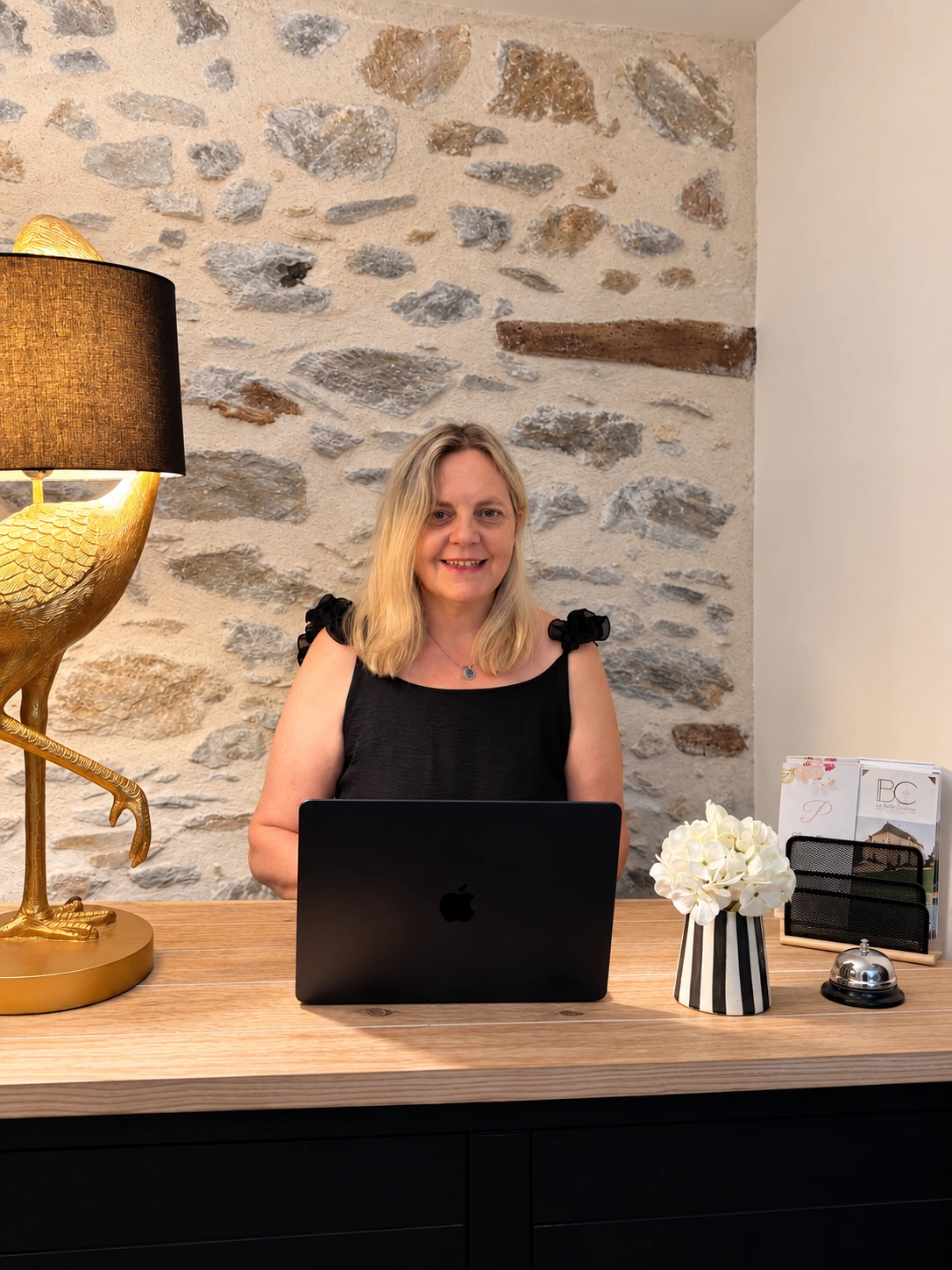 Patricia, votre hôte chez Patou, vous accueille devant le mur en pierre de la maison